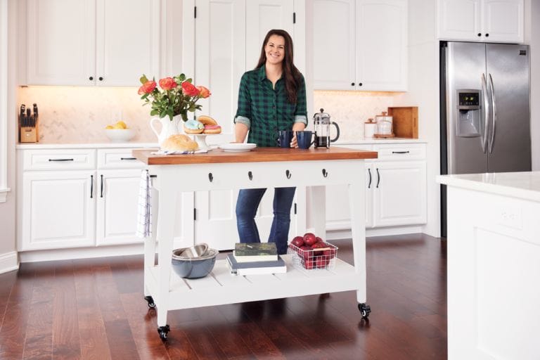 A person stands in a bright kitchen beside a white rolling island with a wooden top. The island holds a bouquet of flowers, loaf of bread, coffee press, and fruit. White cabinets and stainless steel appliances complete the scene.
