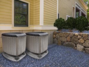 Two heat pumps on the exterior of a yellow home near a stone wall.