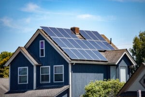 A dark blue large house with white window panels, a grey paneled roof with solar panels, and green trees