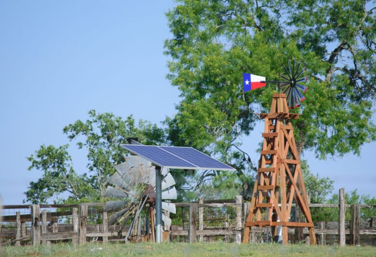 Solar Water well with Texas Windmill, in front of green trees and blue sky. A Texas solar panel sits beside the windmill.